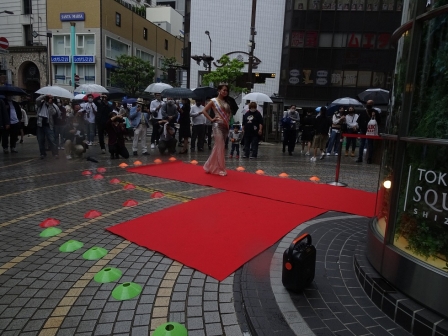 一人のモデルが施設正面入り口前に敷かれた赤いカーペットの上を歩く様子を、観客が見つめている。
