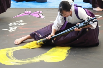 紫色の袴をはいた女子高生が、太く大きな筆に黄色の墨汁をつけて、全身を使って大きな文字を書いている。